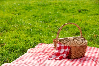 Photo of One picnic wicker basket with checkered napkin and blanket on green grass. Space for text