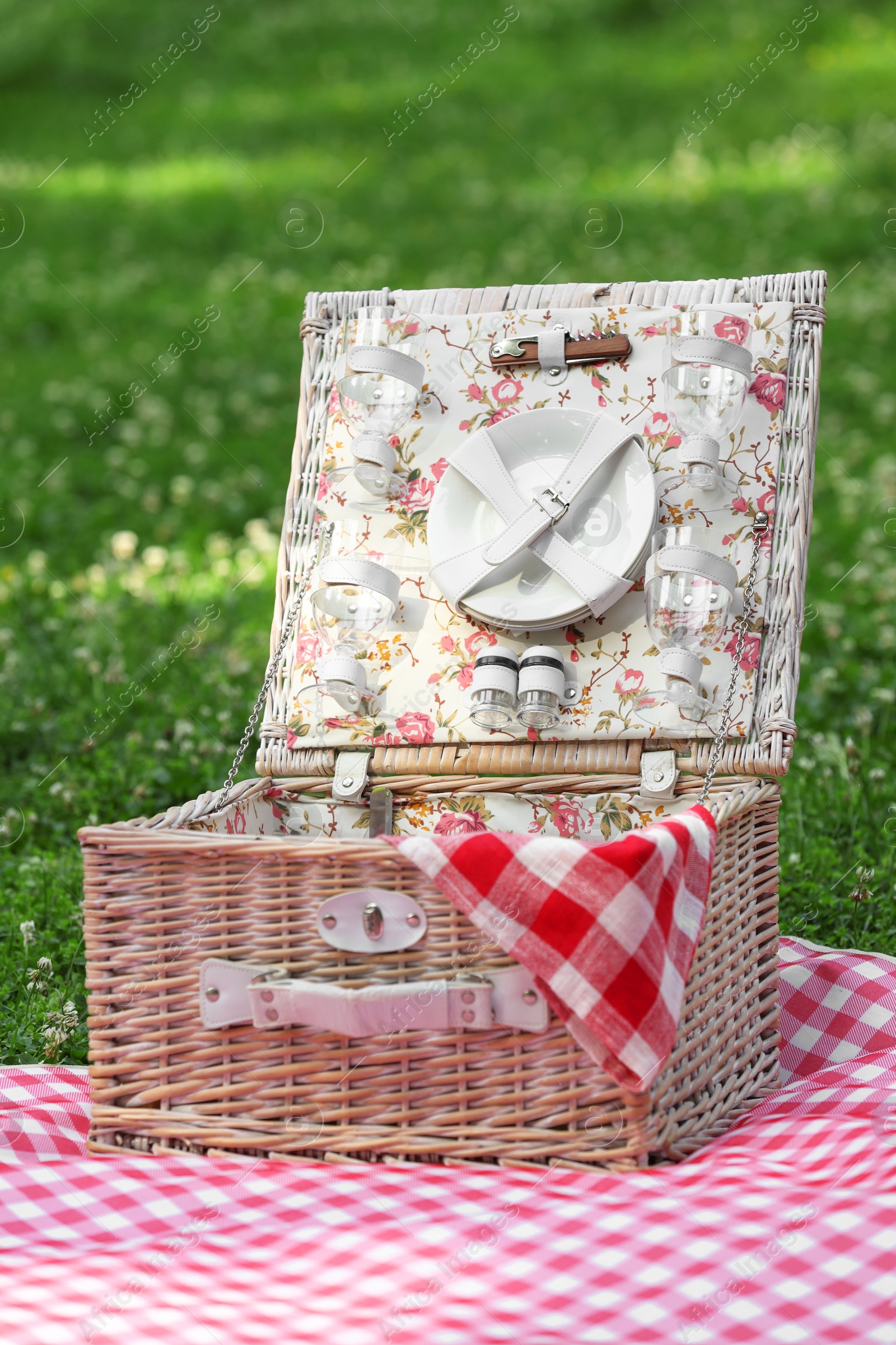 Photo of One picnic wicker basket with checkered napkin, tableware and blanket on green grass