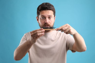 Sad man holding brush with lost hair on light blue background. Alopecia problem