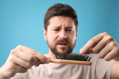 Sad man taking his lost hair from brush on light blue background, selective focus. Alopecia problem