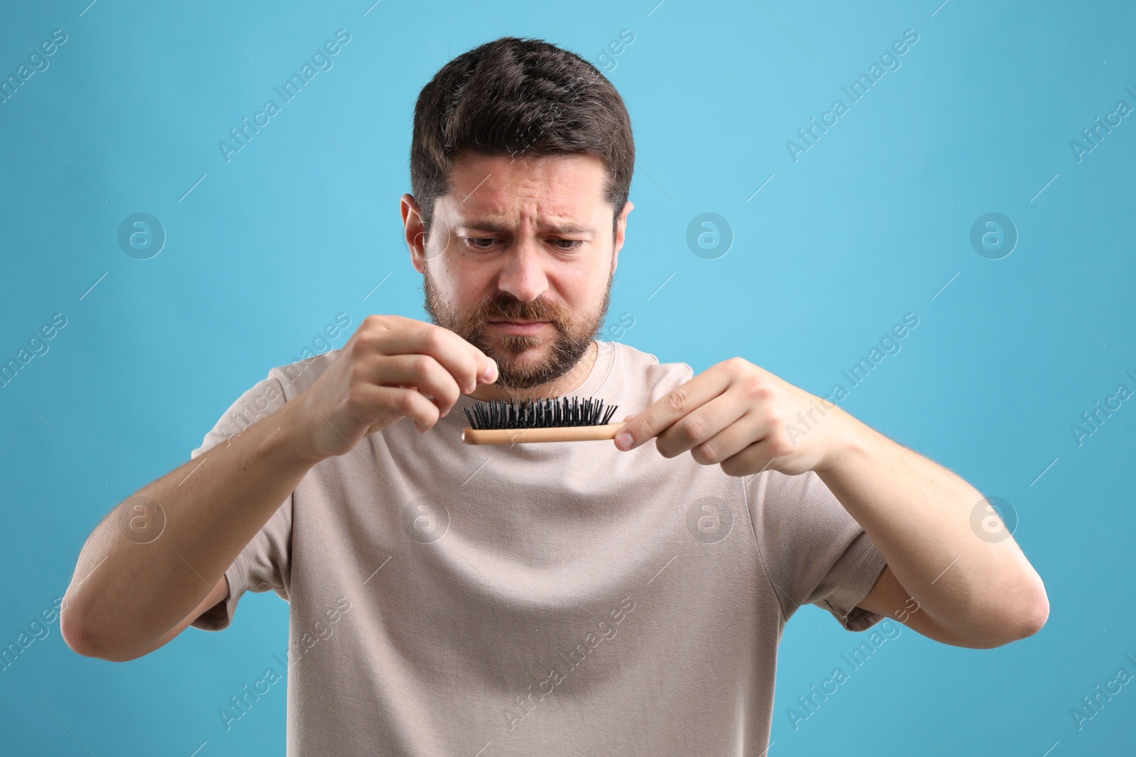 Photo of Sad man taking his lost hair from brush on light blue background. Alopecia problem