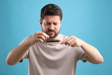 Sad man taking his lost hair from brush on light blue background. Alopecia problem