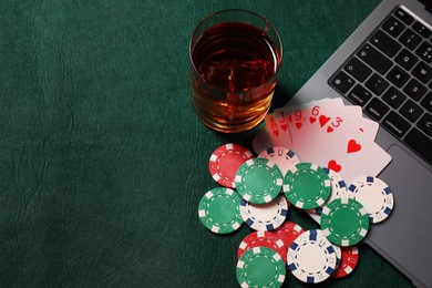 Photo of Poker chips, laptop, playing cards and glass of alcohol drink on green table, flat lay. Online game