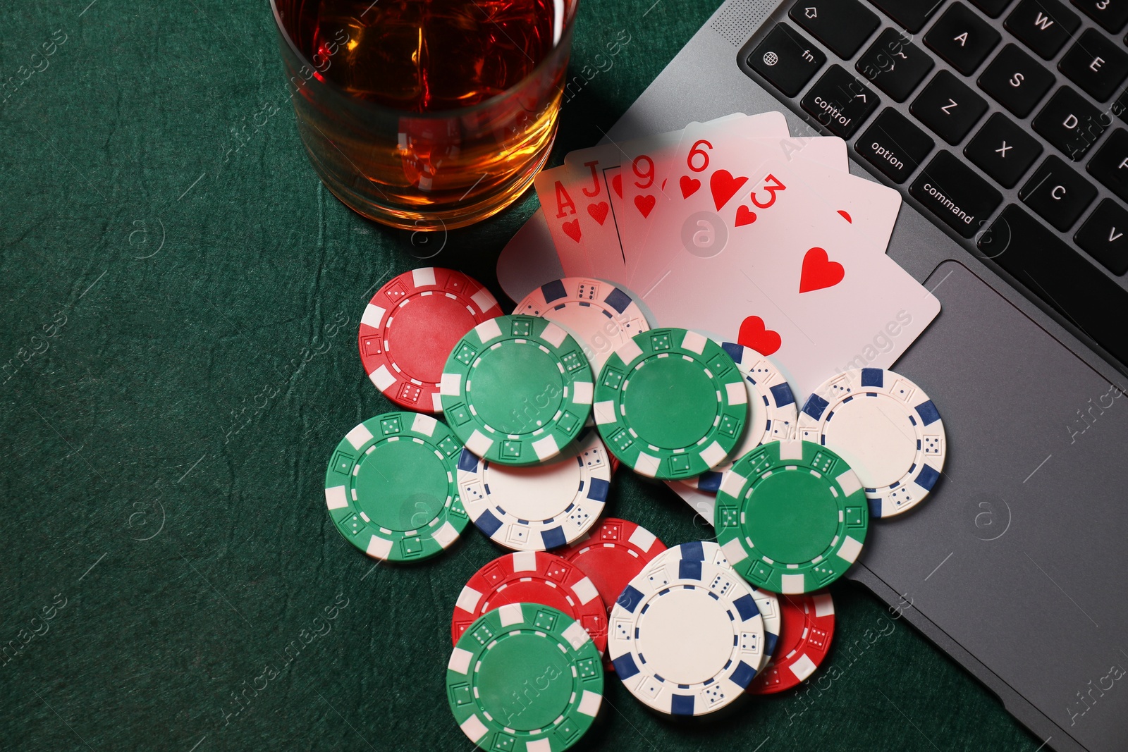 Photo of Poker chips, laptop, playing cards and glass of alcohol drink on green table, flat lay. Online game