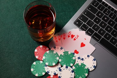 Photo of Poker chips, laptop, playing cards and glass of alcohol drink on green table, flat lay. Online game