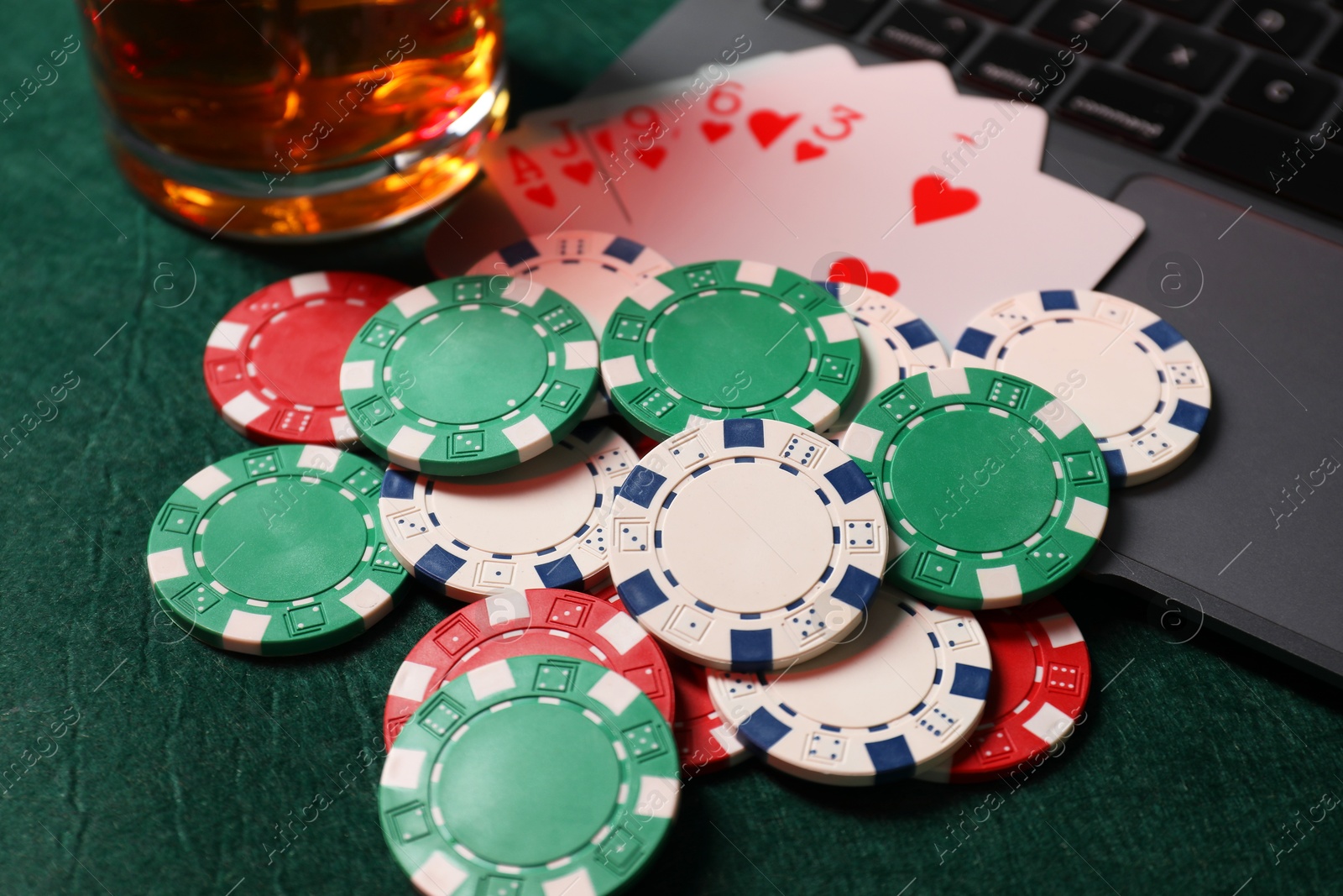 Photo of Poker chips, laptop, playing cards and glass of alcohol drink on green table, closeup. Online game