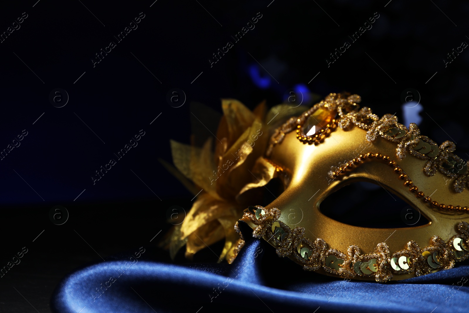 Photo of Beautiful golden carnival mask on blue fabric in darkness, closeup