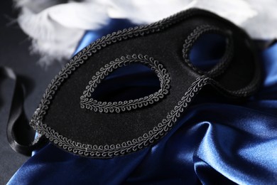 Photo of Beautiful carnival mask, white feathers and blue silk fabric on black background, closeup