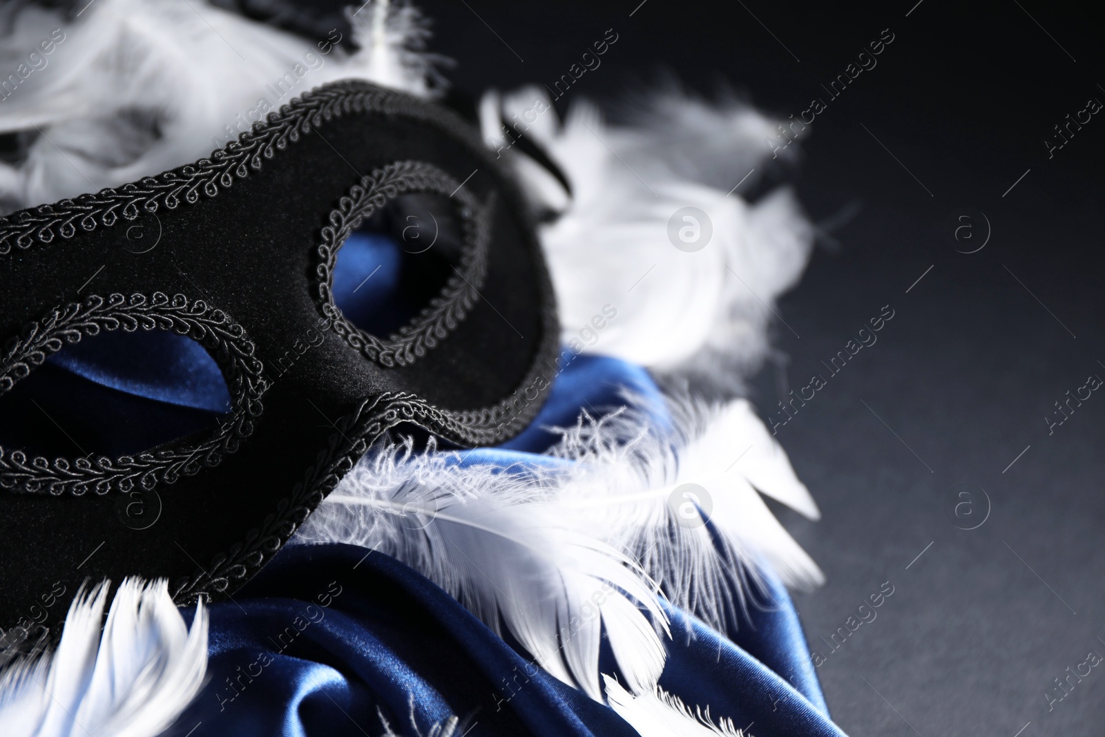 Photo of Beautiful carnival mask, white feathers and blue silk fabric on black background, closeup