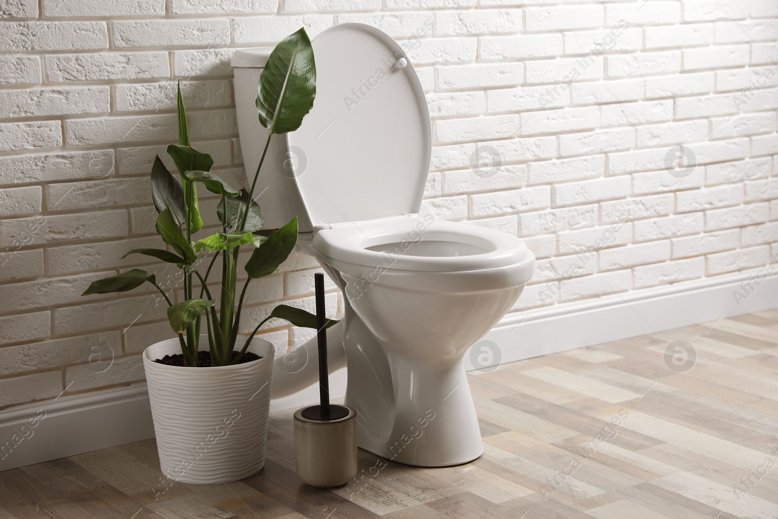 Photo of Ceramic toilet bowl, brush and houseplant near white brick wall in restroom