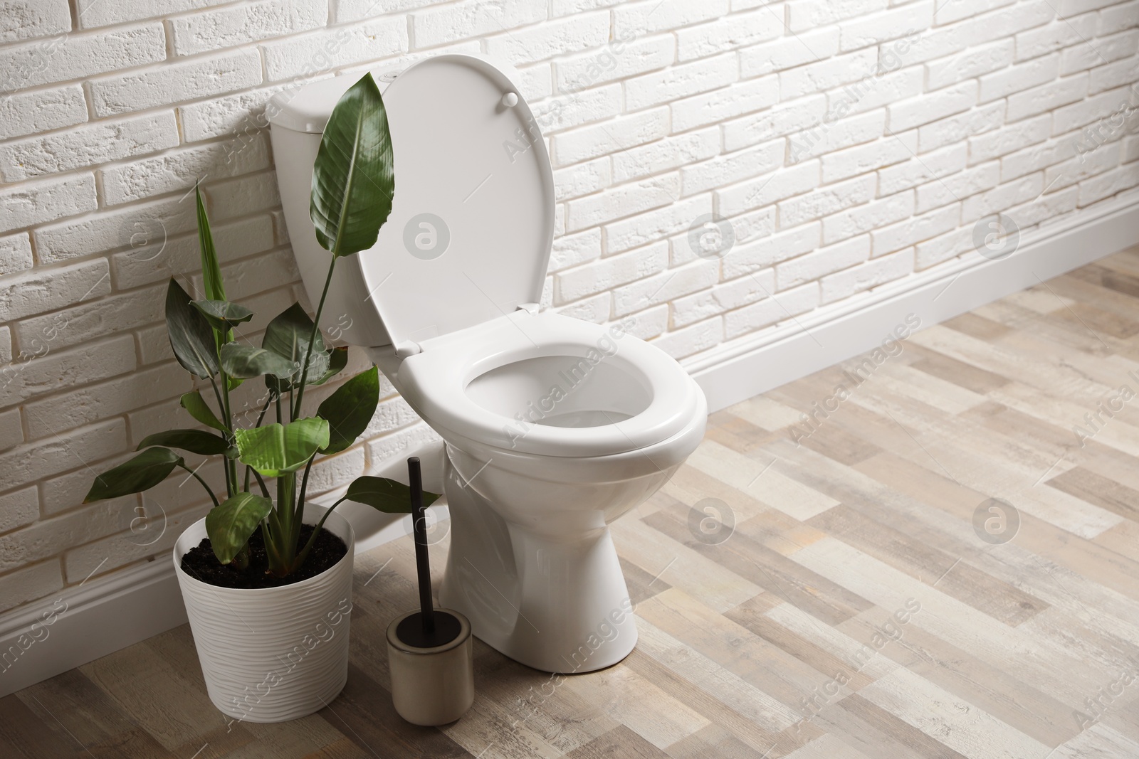 Photo of Ceramic toilet bowl, brush and houseplant near white brick wall in restroom. Space for text