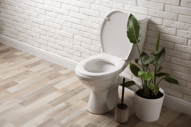 Photo of Ceramic toilet bowl, brush and houseplant near white brick wall in restroom. Space for text