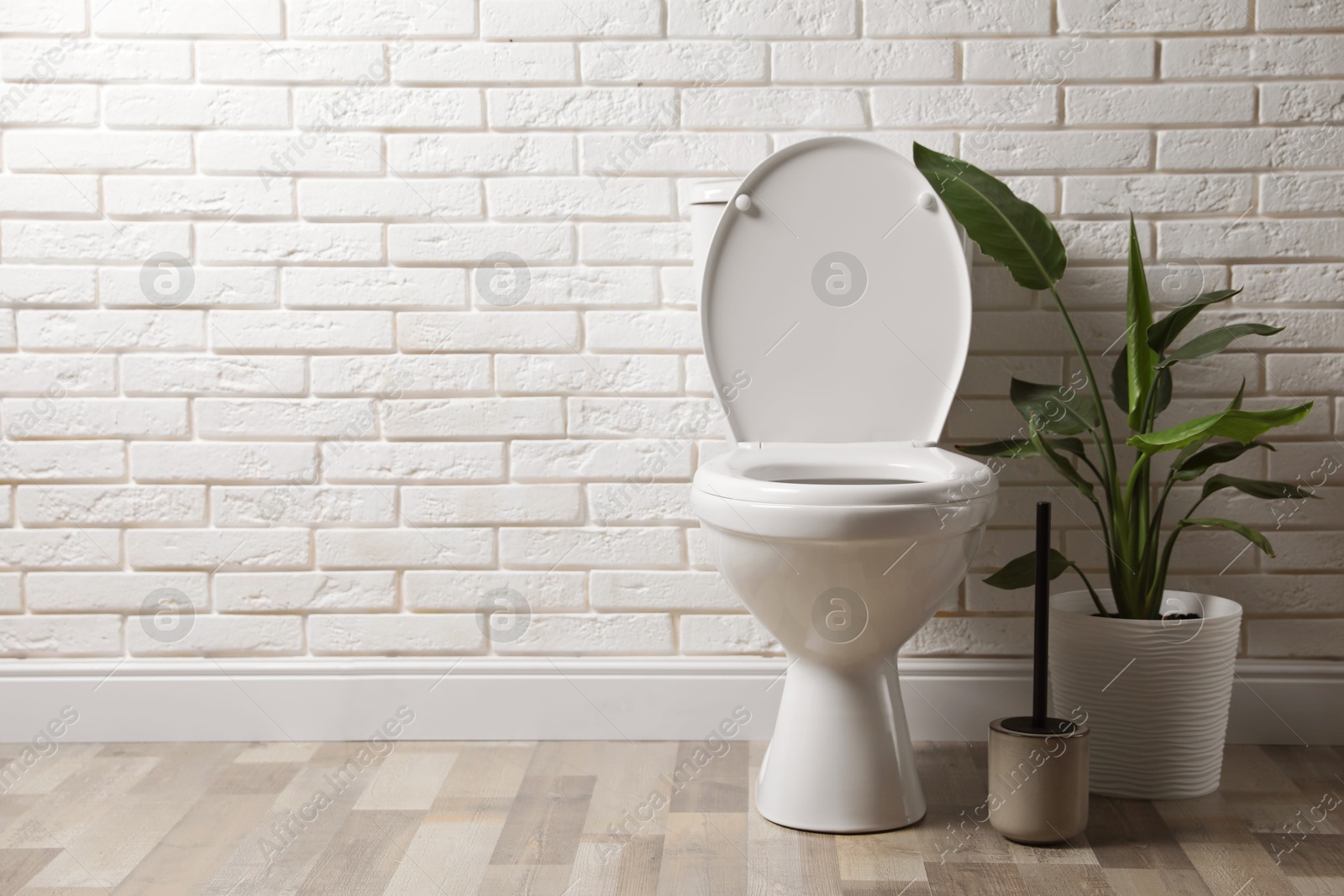 Photo of Ceramic toilet bowl, brush and houseplant near white brick wall in restroom. Space for text