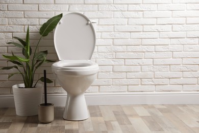 Photo of Ceramic toilet bowl, brush and houseplant near white brick wall in restroom. Space for text