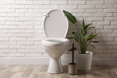 Photo of Ceramic toilet bowl, brush and houseplant near white brick wall in restroom