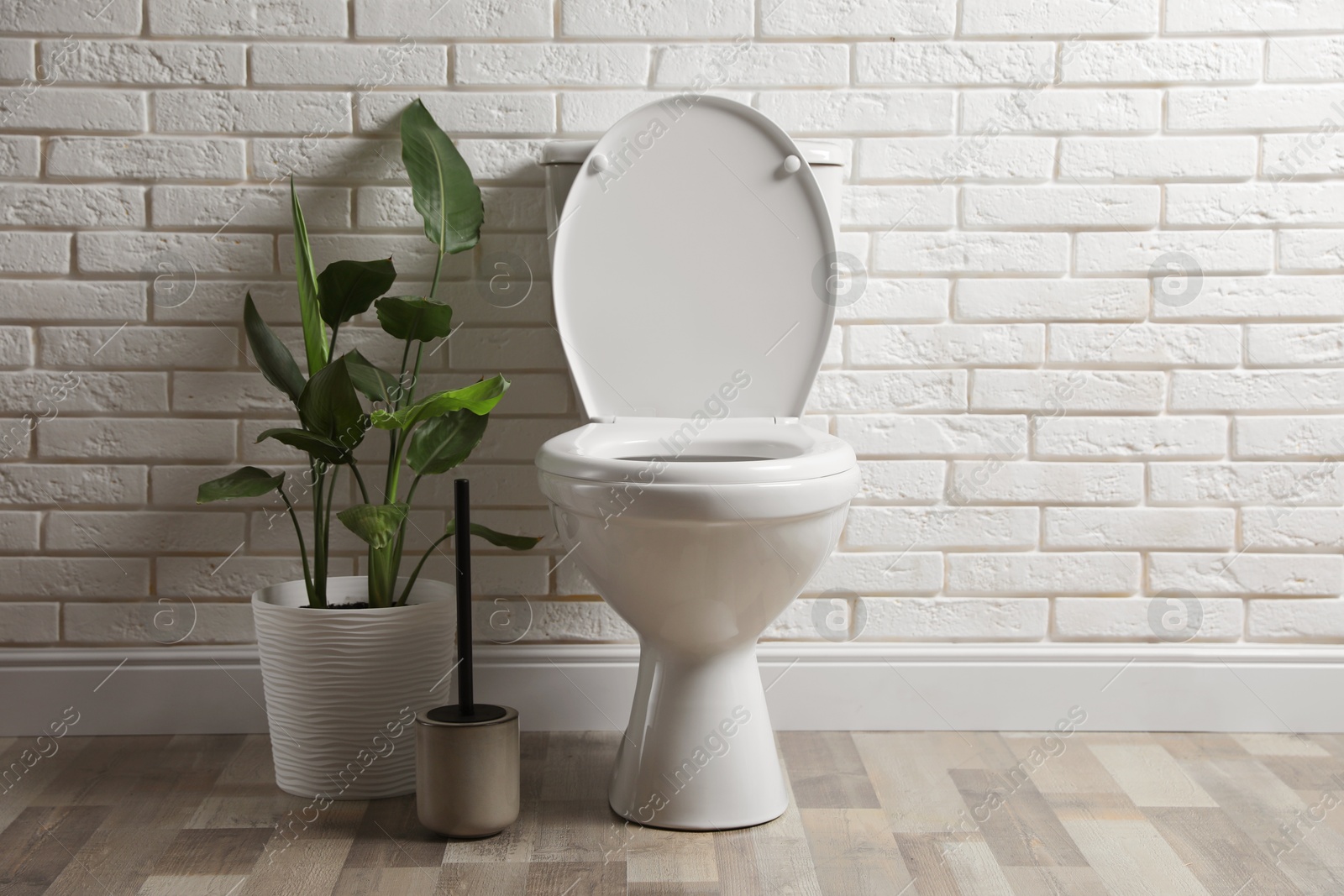 Photo of Ceramic toilet bowl, brush and houseplant near white brick wall in restroom