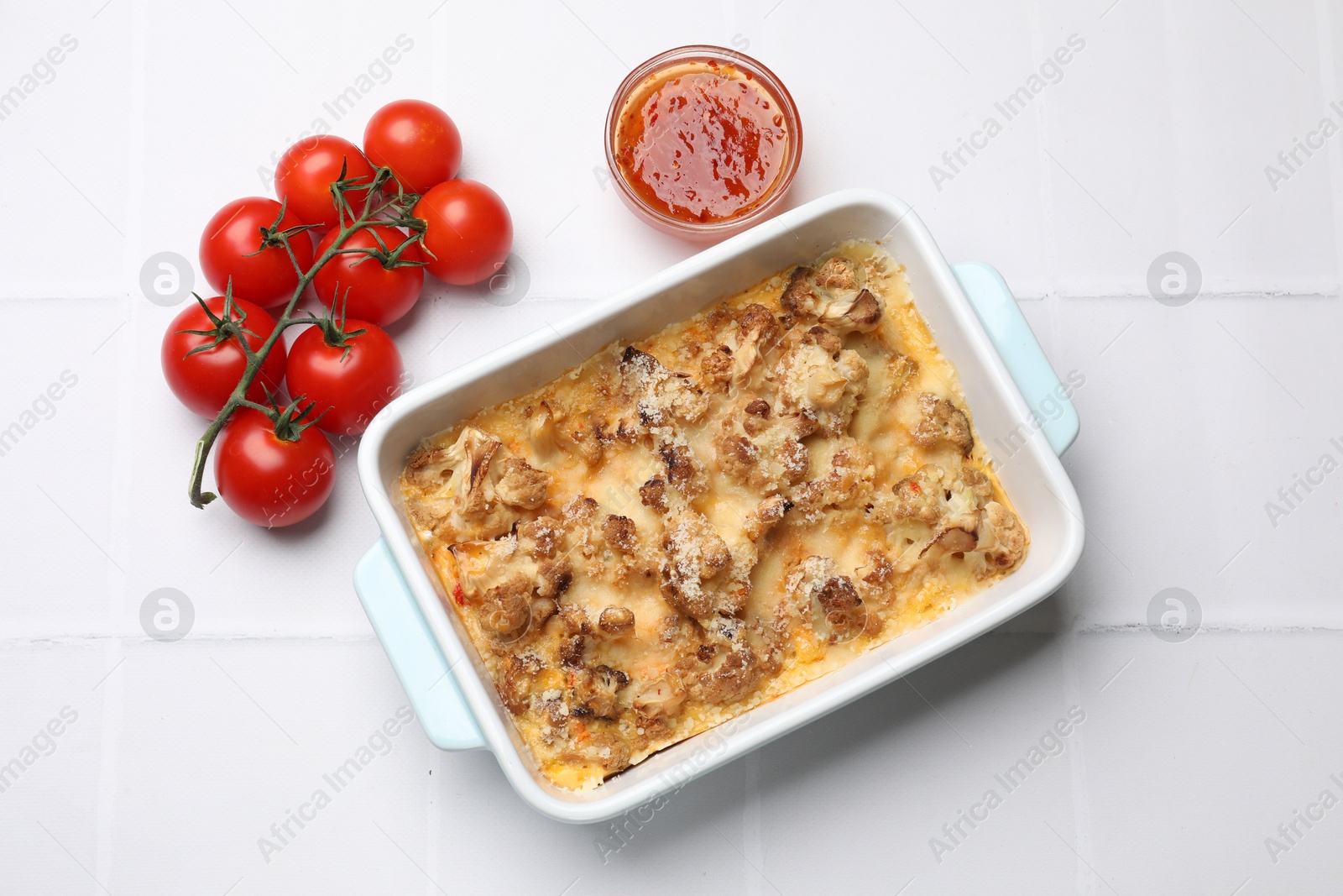 Photo of Tasty baked cauliflower, tomatoes and sauce on white tiled table, flat lay