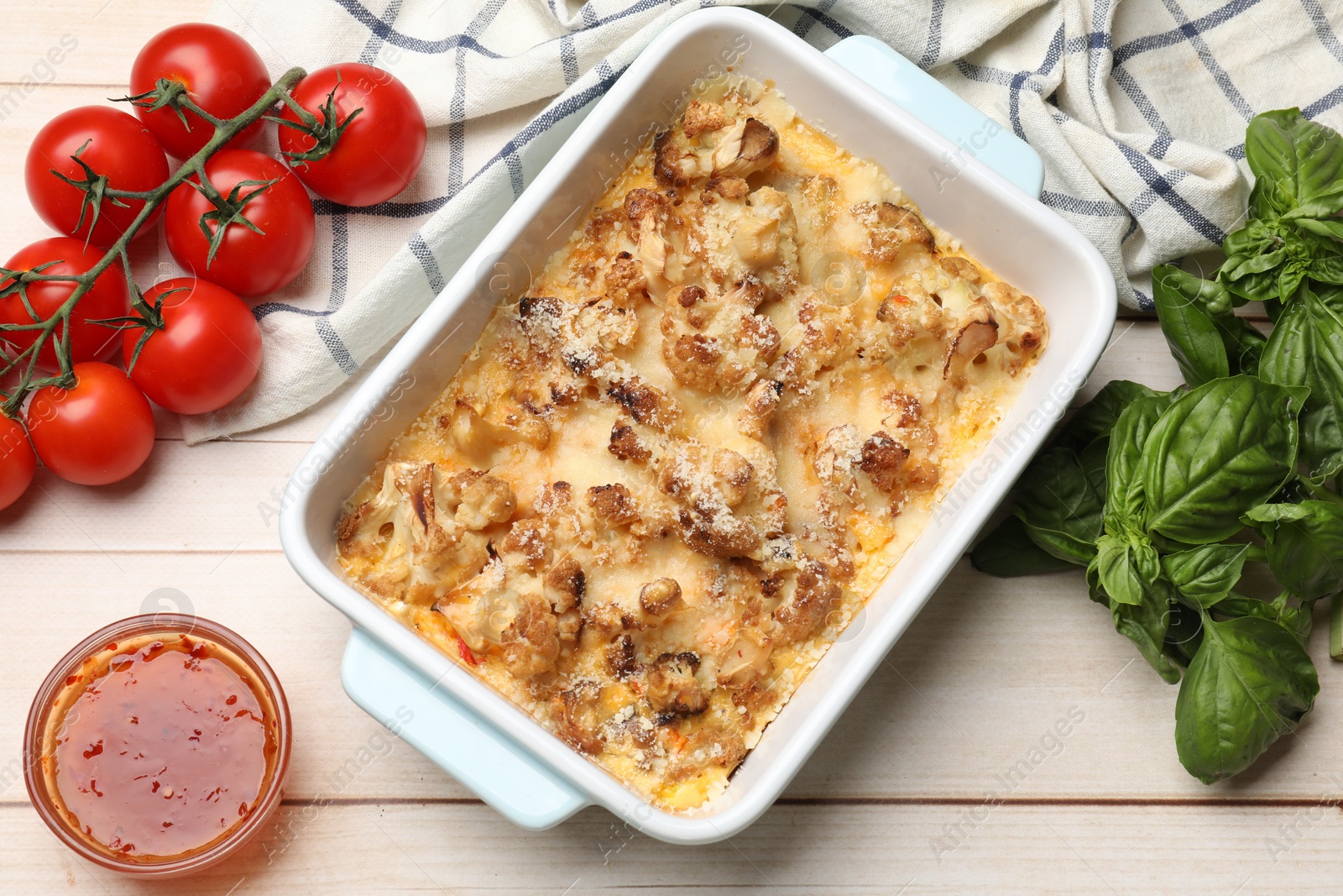 Photo of Tasty baked cauliflower in baking dish, tomatoes, basil and sauce on light wooden table, flat lay