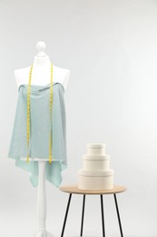 Mannequin with measuring tape, fabric and boxes on side table against light grey background. Creating new outfit