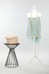 Photo of Mannequin with measuring tape, fabric and box on side table against light grey background. Creating new outfit