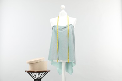 Photo of Mannequin with measuring tape, fabric and box on side table against light grey background. Creating new outfit