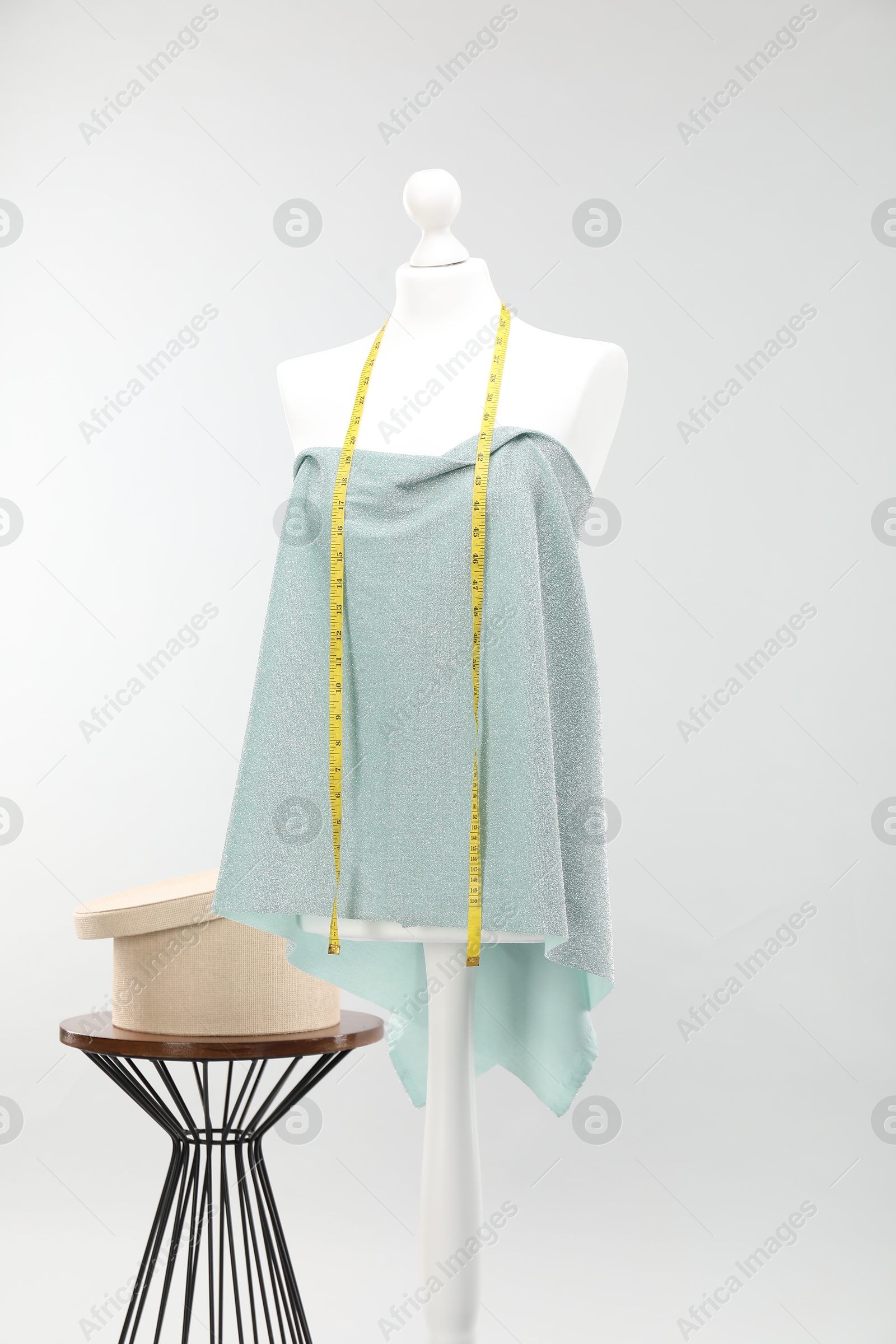 Photo of Mannequin with measuring tape, fabric and box on side table against light grey background. Creating new outfit