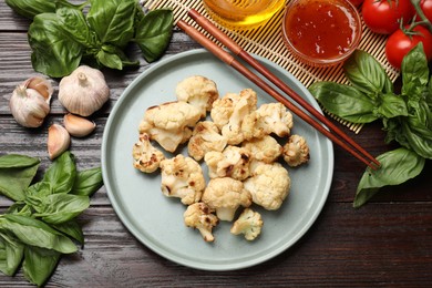 Plate with baked cauliflower, products, chopsticks and sauce on wooden table, flat lay