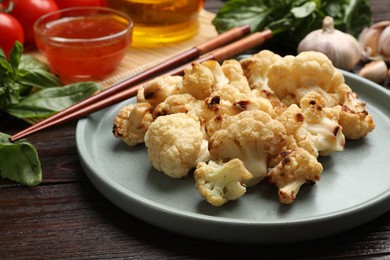 Photo of Plate with baked cauliflower, products, chopsticks and sauce on wooden table, closeup