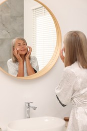 Photo of Senior woman applying face cream near mirror at home