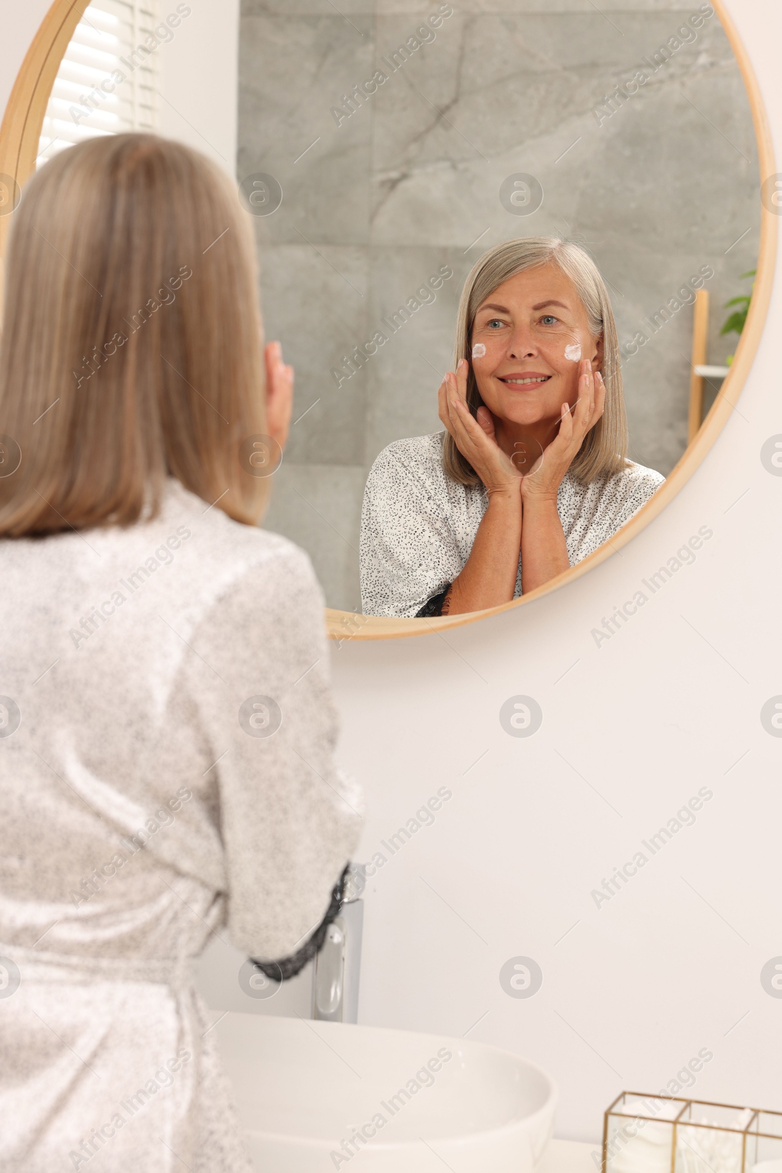 Photo of Senior woman applying face cream near mirror at home