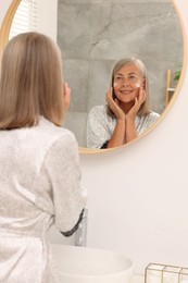 Senior woman applying face cream near mirror at home