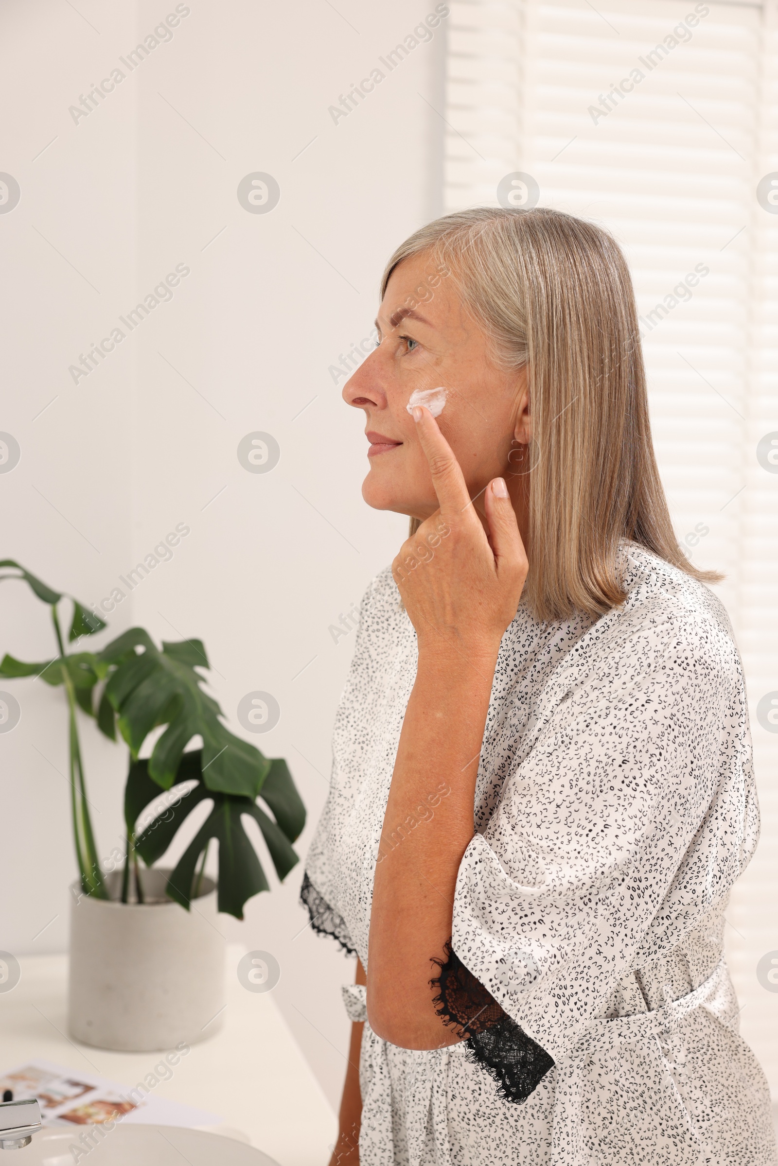 Photo of Senior woman applying face cream near mirror at home