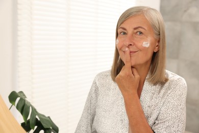 Senior woman applying face cream near mirror at home