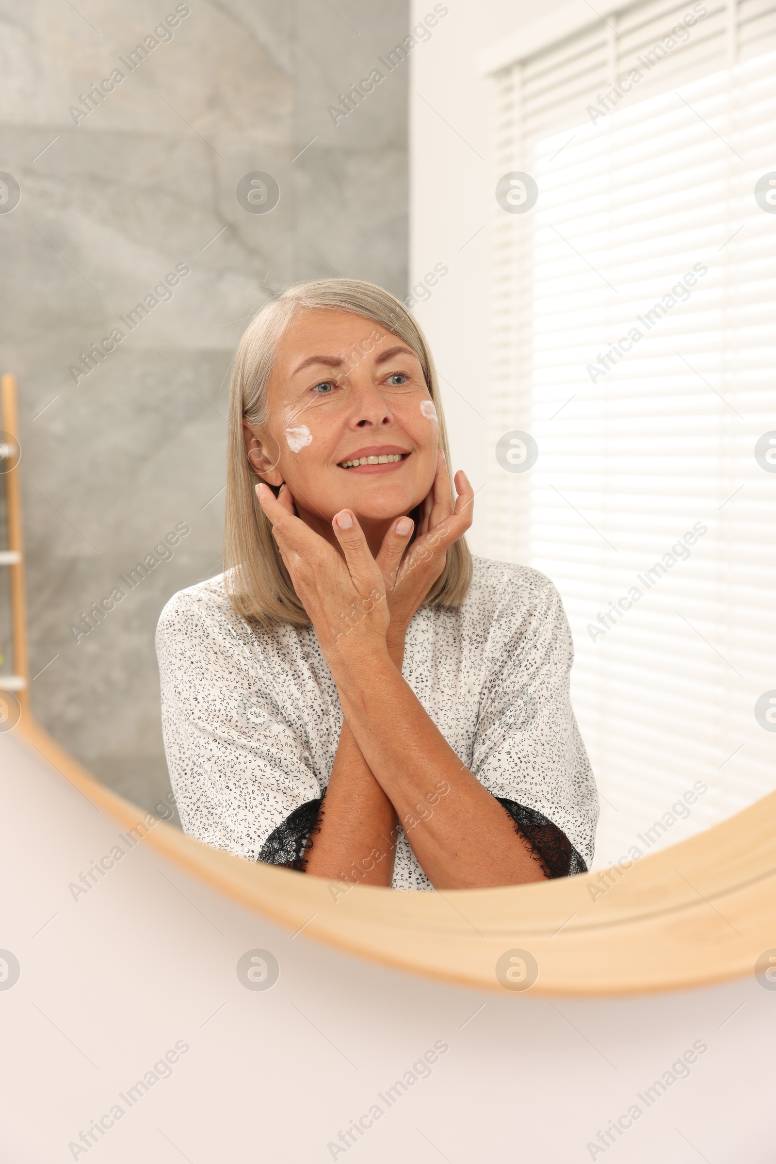 Photo of Senior woman applying face cream near mirror at home