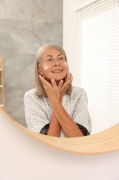 Senior woman applying face cream near mirror at home