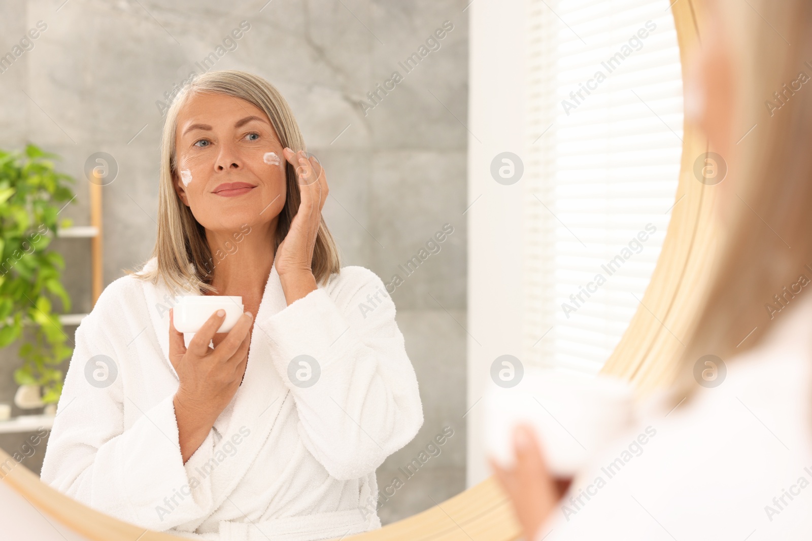 Photo of Senior woman applying face cream near mirror at home