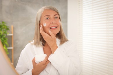 Photo of Senior woman applying face cream near mirror at home