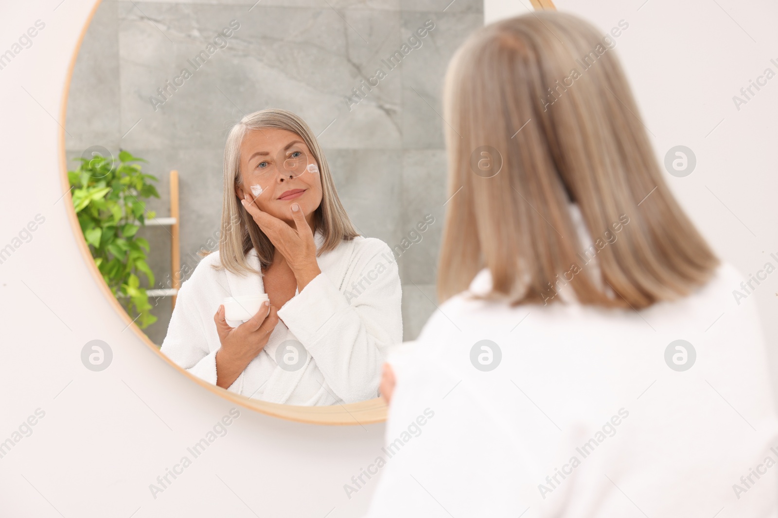 Photo of Senior woman applying face cream near mirror at home