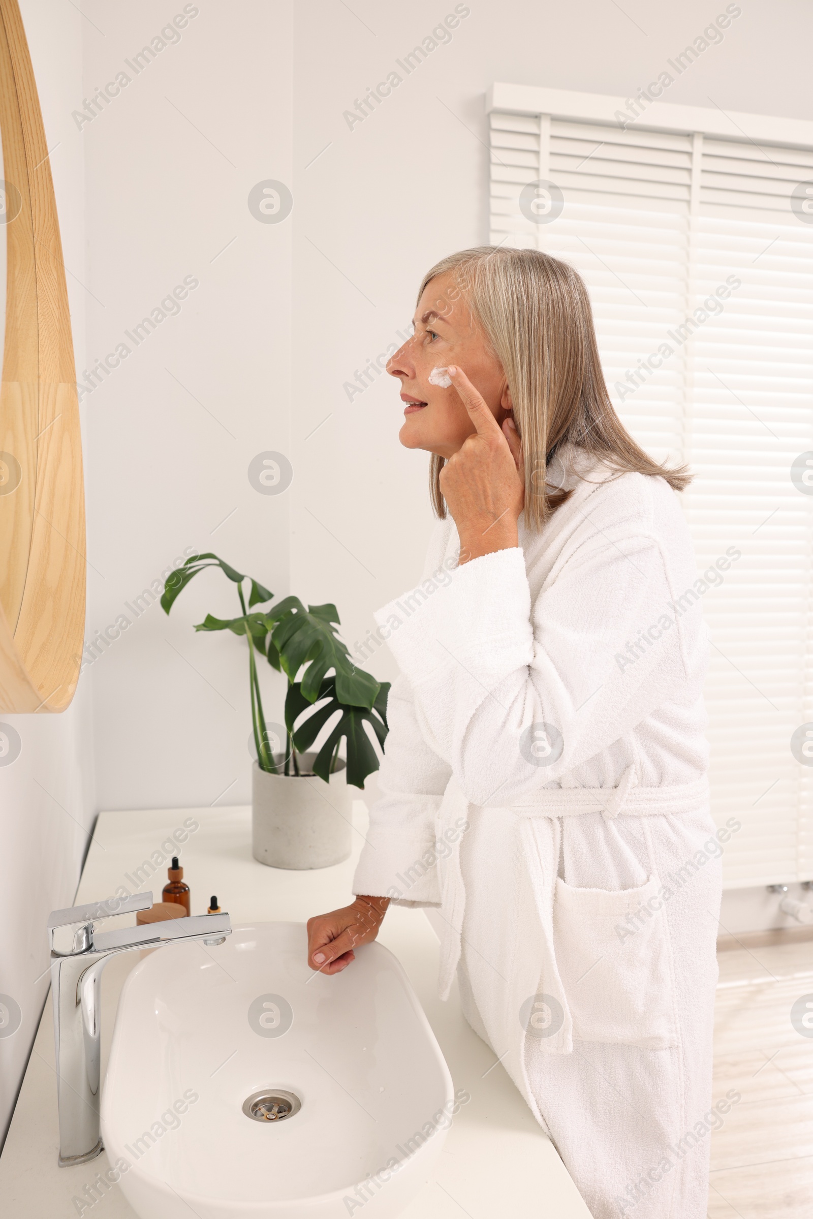 Photo of Senior woman applying face cream near mirror at home