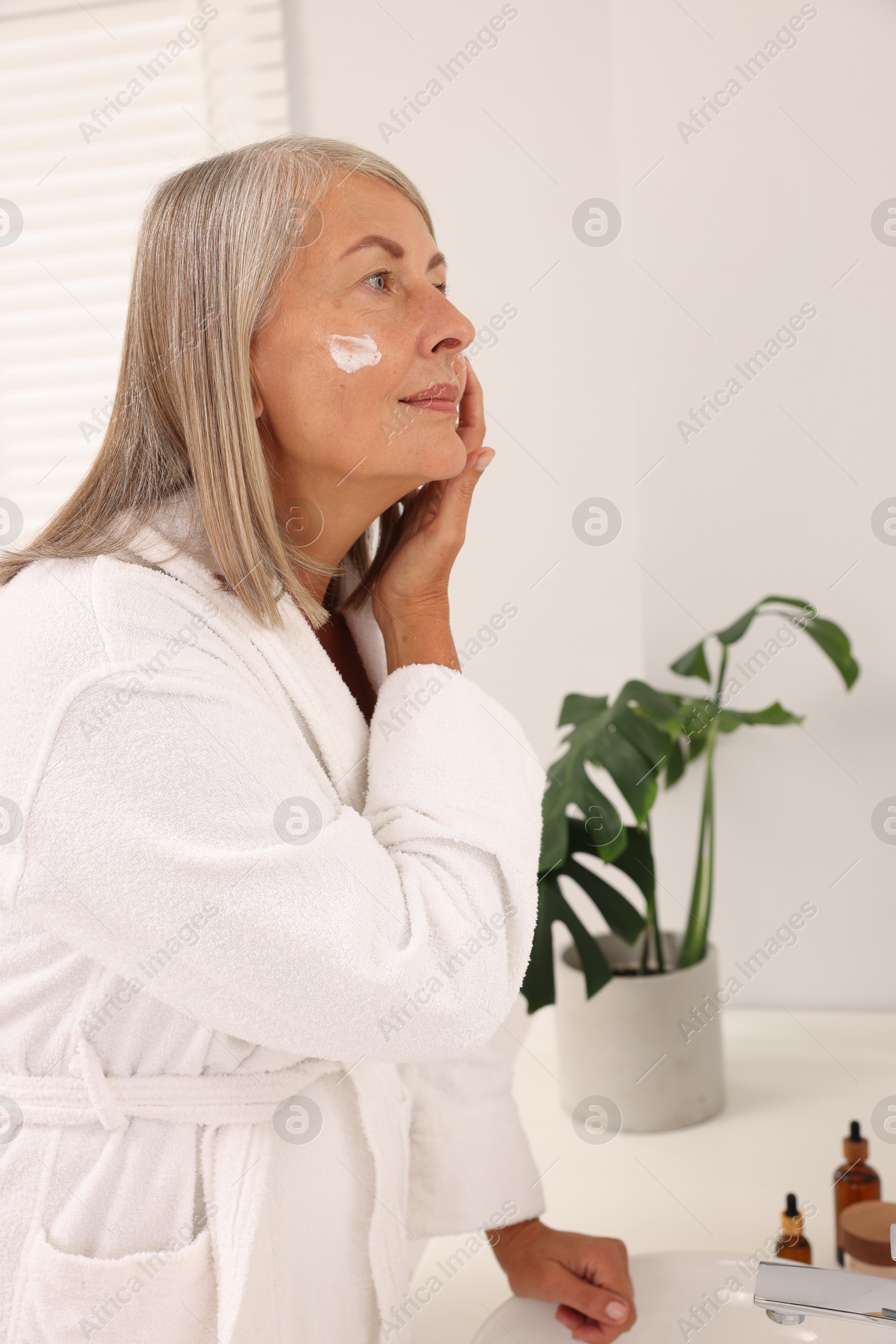 Photo of Senior woman applying face cream near mirror at home