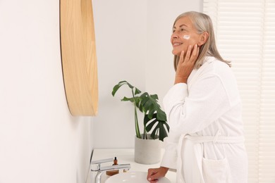 Senior woman applying face cream near mirror at home