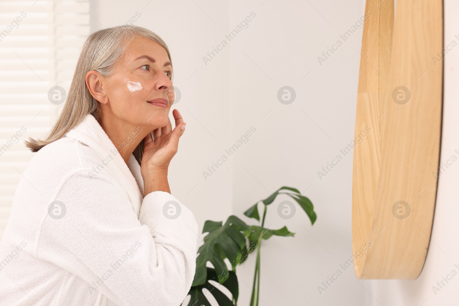 Photo of Senior woman applying face cream near mirror at home