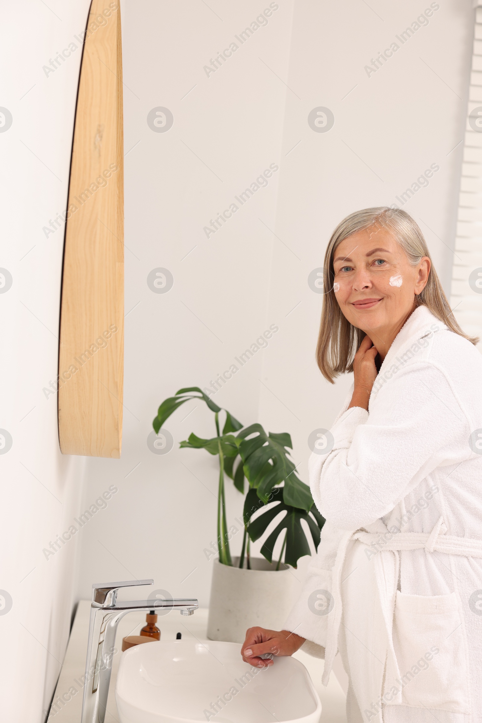 Photo of Senior woman applying face cream near mirror at home