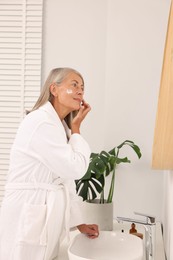 Photo of Senior woman applying face cream near mirror at home