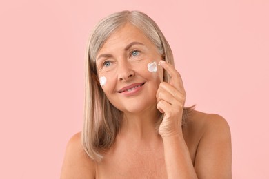 Senior woman applying face cream on pink background