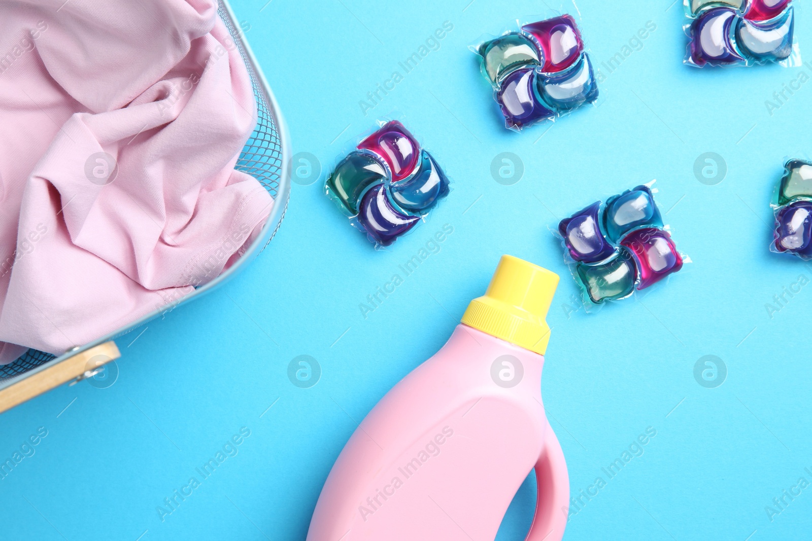 Photo of Basket with clothes, bottle of fabric softener and laundry detergent pods on blue background, flat lay