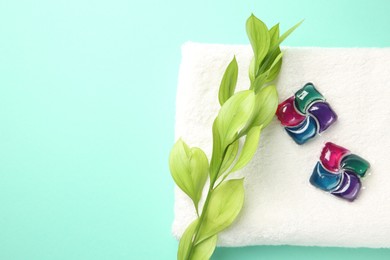 Photo of Laundry detergent pods, towel and green branch on turquoise background, flat lay. Space for text