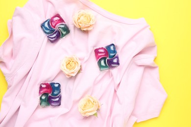 Photo of Laundry detergent pods, t-shirt and flowers on yellow background, flat lay