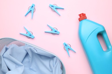 Photo of Basket with clothes, bottle laundry detergent and clothespins on pink background, flat lay