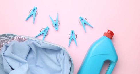 Photo of Basket with clothes, bottle laundry detergent and clothespins on pink background, flat lay